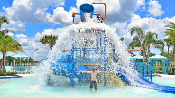 Zona de juegos acuáticos con un niño en el agua, rodeado de palmeras y un cielo despejado.