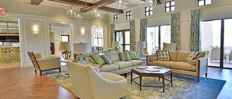 A spacious living room with wooden flooring, a large sofa set, and a coffee table. There are multiple windows with curtains and a chandelier.