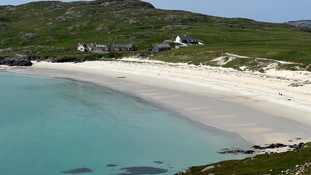 Une zone côtière avec une plage de sable, des eaux turquoise et limpides, et un groupe de maisons sur une colline verdoyante.