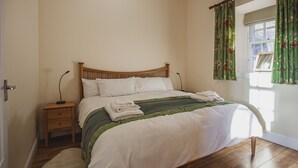 Un lit bien fait, recouvert d’une couvre-lit verte et blanche, une table de chevet en bois avec une lampe, et une fenêtre ornée de rideaux à motifs floraux.