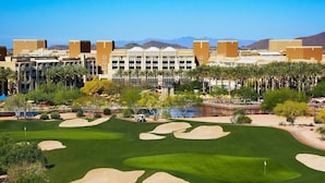 A golf course with bunkers, a water body, and a hotel with multiple floors and a desert landscape in the background. - Super Bowl Week! (Phoenix)