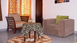 Een kamer met een bank, een stoel, een kleintje tafeltje met een tafelkleed met patroon, een kleurrijk gordijn en een deur.