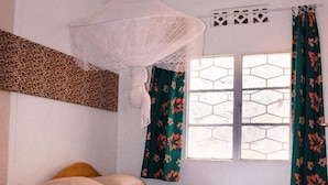 Een slaapkamer met een bed, een nachtkastje, een raam met patroon gordijnen en een hangend klamboe.