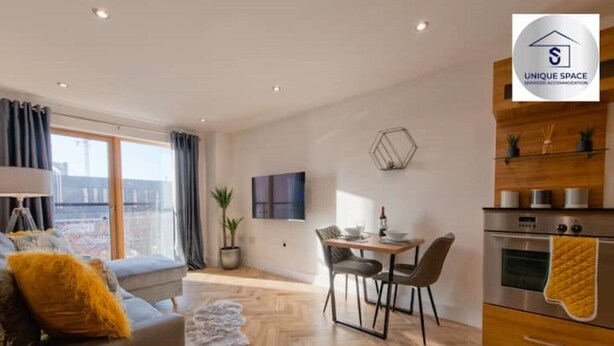 A modern living room with a sofa, a dining table with chairs, a potted plant, and a kitchen area with an oven and a countertop.