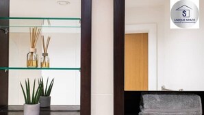 A bathroom with a glass shelf holding reed diffusers and potted plants, a wooden vanity with a towel, and a logo for Unique Space.