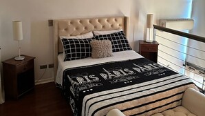 A bedroom with a tufted headboard, a black and white striped bedspread, a bedside table with a lamp, and a window with a view. - Central Suites (Santiago)