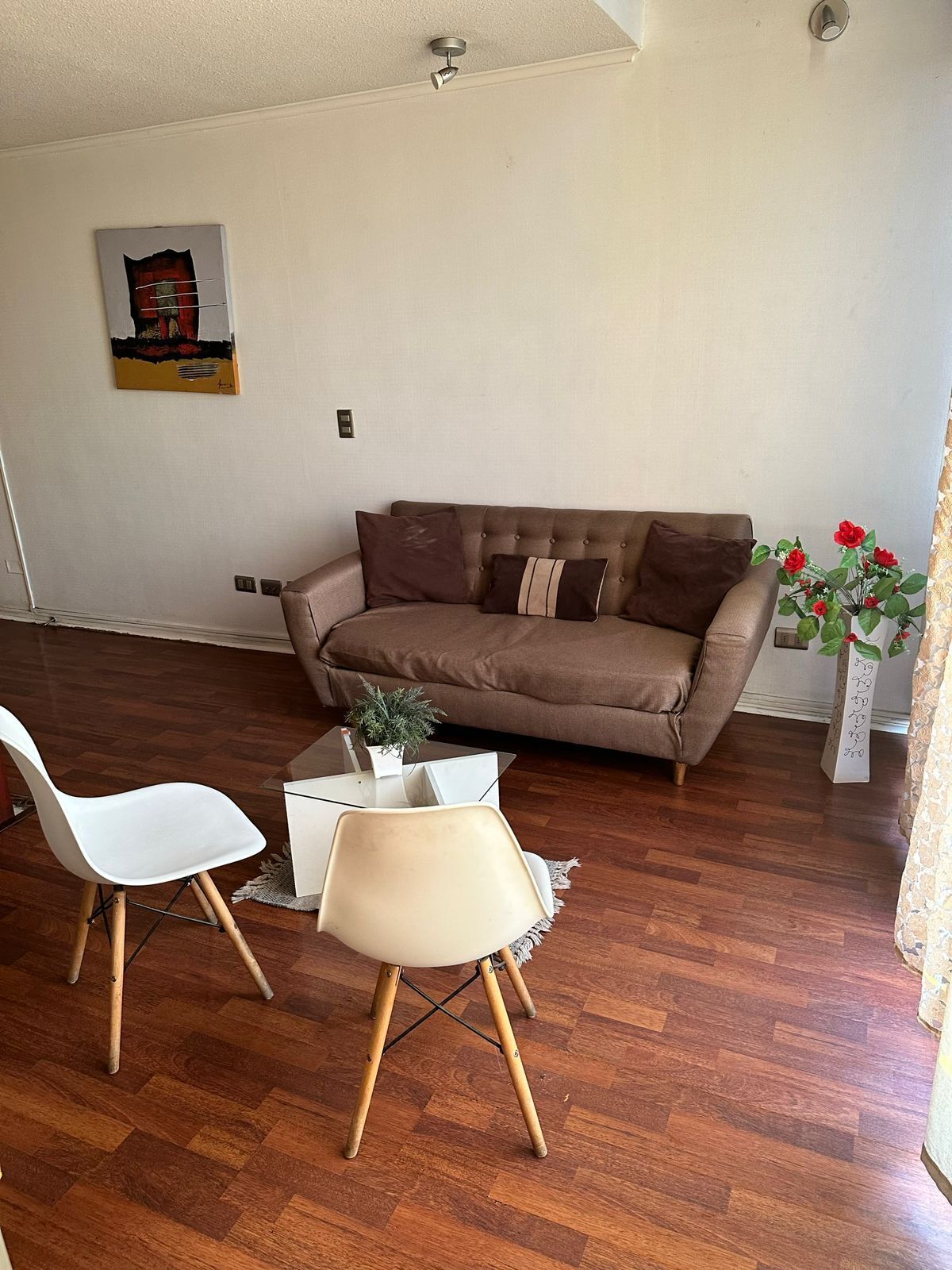 Una sala de estar con un sofá marrón, dos sillas blancas, una pequeña mesa de centro y una planta en maceta.