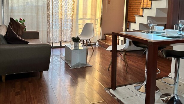 A modern living room with a wooden floor, a sofa, a dining table, and a staircase with a brick wall. - Central Suites (Santiago)