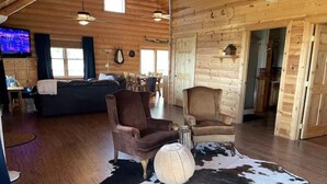 A spacious living area with a wooden floor, a large black and white cowhide rug, two leather armchairs, and a television mounted on the wall.