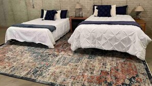 A room with two beds, each with a white bedspread and dark blue pillows. There is a patterned rug on the floor.
