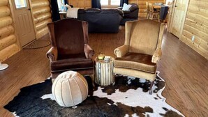 A log cabin interior with a living room featuring two armchairs, a round ottoman, and a cowhide rug.