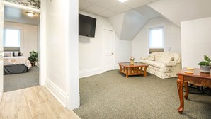 Une chambre spacieuse aménagée en mezzanine, dotée d’une grande fenêtre, d’un téléviseur à écran plat fixé au mur, d’une table basse en bois, d’un canapé et d’une petite table sur laquelle est posée une plante en pot.