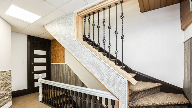 Un escalier avec une rampe en bois, des balustrades noires et des marches recouvertes d’une moquette beige.