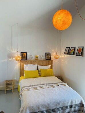 A neatly arranged bedroom with a wooden headboard, a bed with white and yellow bedding, and a hanging orange pendant light.