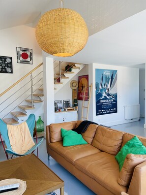 A modern living room with a brown leather sofa, green and beige cushions, a wicker pendant light, a staircase with wooden steps, and a poster of a sailing boat.