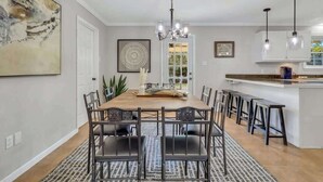 Ein moderner Essbereich mit Holztisch und schwarzen Stühlen, einer Kristallleuchter und einer Küchenzeile mit weißen Schränken und Pendelleuchten.