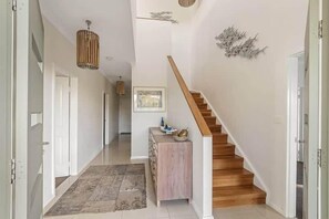 A hallway with a wooden staircase, a white wall with a decorative piece, and a hanging light fixture.