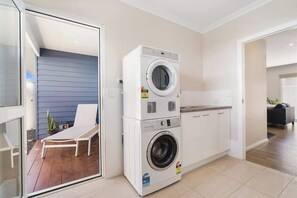 A laundry room with stacked washers and dryers, a chair, and a view into a living area with a sofa and a plant.