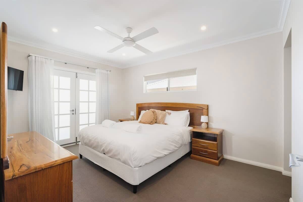 A bedroom with a bed, bedside tables, a ceiling fan, a TV mounted on the wall, and a window with curtains.
