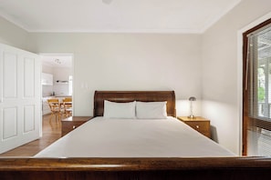 A bedroom with a wooden bed, white bedding, a nightstand with a lamp, a wooden dresser, and a view through a window with blinds.