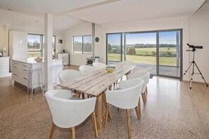 A spacious dining area with a wooden table and white chairs, large windows with a view of the countryside, and a telescope on a tripod. - Margaret River Farmhouse: Serene Bushland Getaway (Rosa Glen)