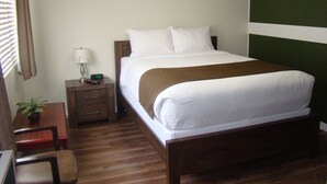 A hotel room with a single bed, a nightstand with a lamp and a clock, a wooden chair, and a small table with a plant. - Paddock Inn (Coleman)
