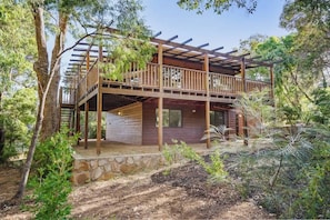 A two-story wooden cabin with a balcony, surrounded by trees and a stone foundation.