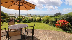 Un patio con mesa y sillas, un paraguas grande y vistas al mar y a una colina con un castillo.