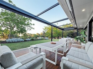 An outdoor patio with a wooden deck, white furniture, and a glass enclosure.