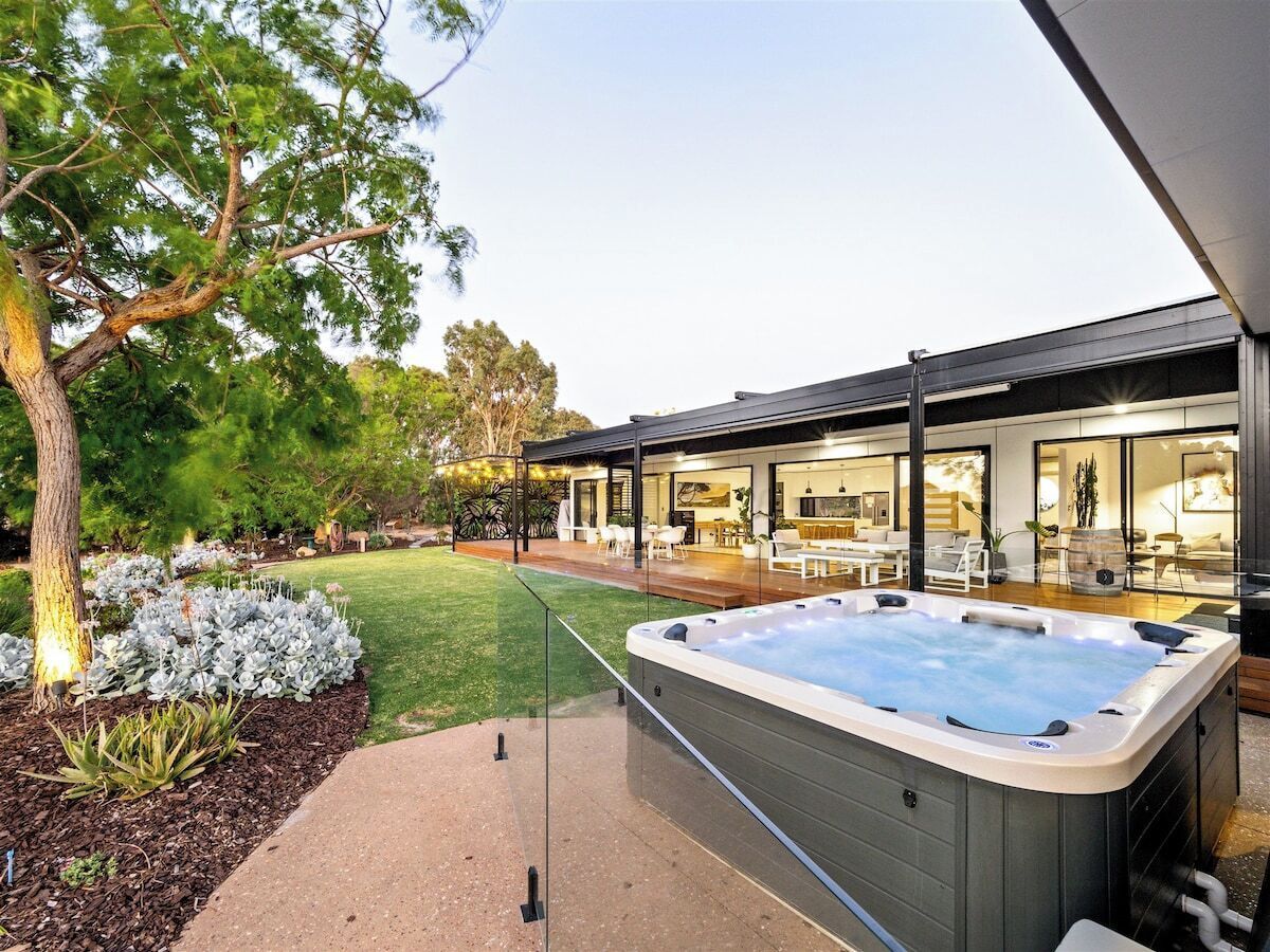 A modern outdoor hot tub area with a glass enclosure, surrounded by a well-maintained garden and a patio with outdoor furniture.