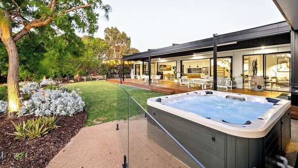 A modern outdoor hot tub area with a glass enclosure, surrounded by a well-maintained garden and a patio with outdoor furniture. - Seaview Yallingup—Nature & Spa Sanctuary (Yallingup)