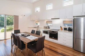 A modern kitchen with white cabinets, a stainless steel refrigerator, and a dining area with a wooden table and chairs. - Villa on the Cape (Dunsborough)