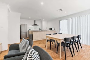 A modern kitchen with a central island, a dining area with a wooden table and chairs, and a sofa with patterned cushions.