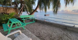 A beach with a green bench and white lounge chairs, palm trees, and a boat in the water.