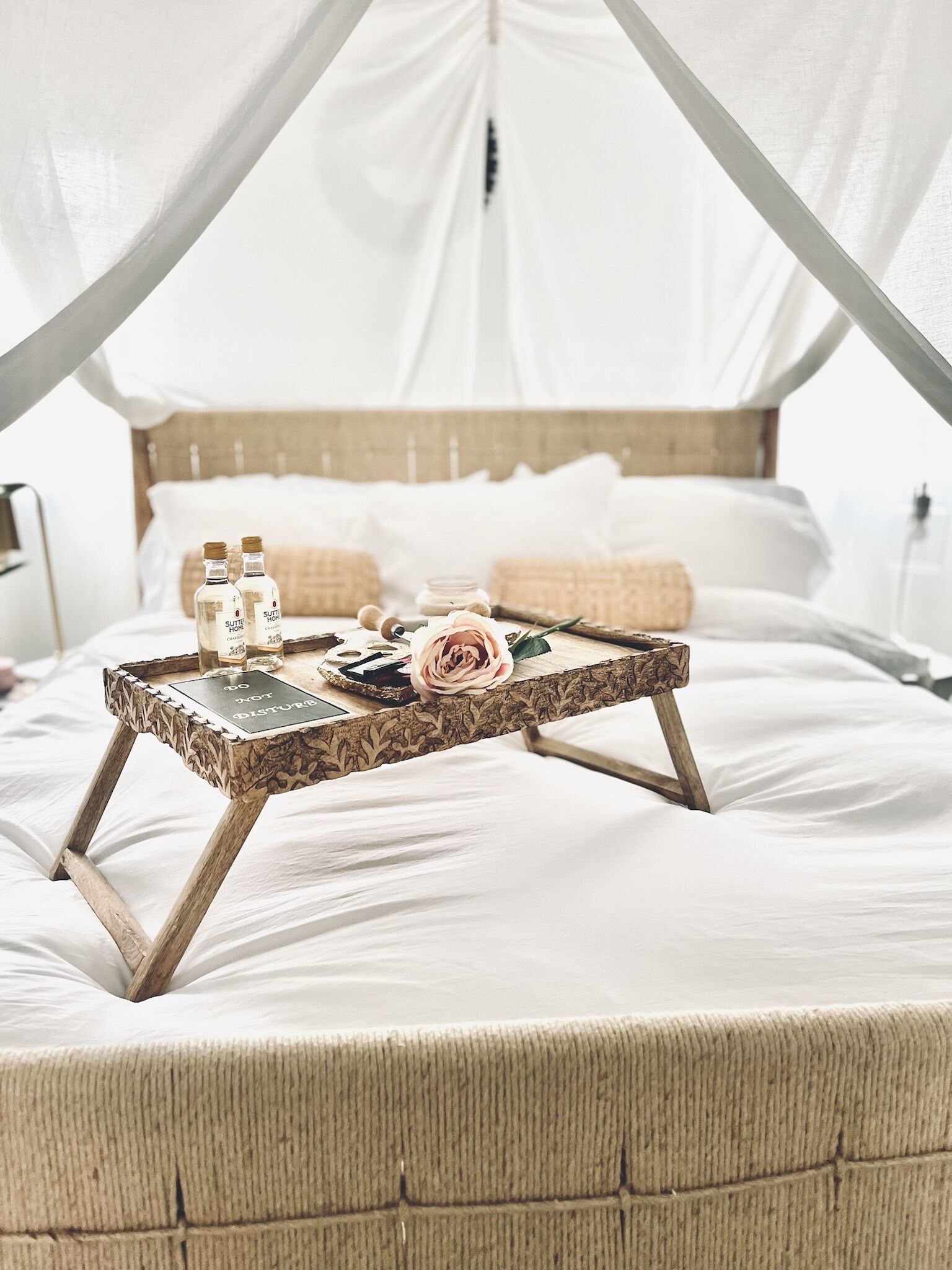 Una cama con dosel, una bandeja de madera con botellas y una rosa, y un almohadón.