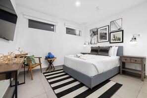 Un dormitorio moderno con una cama grande, un escritorio y una silla. Hay cuadros en la pared y una ventana con persianas.