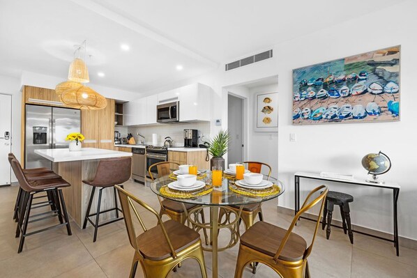 Una cocina moderna con armarios blancos, una isla central y un comedor con mesa de vidrio y sillas doradas.