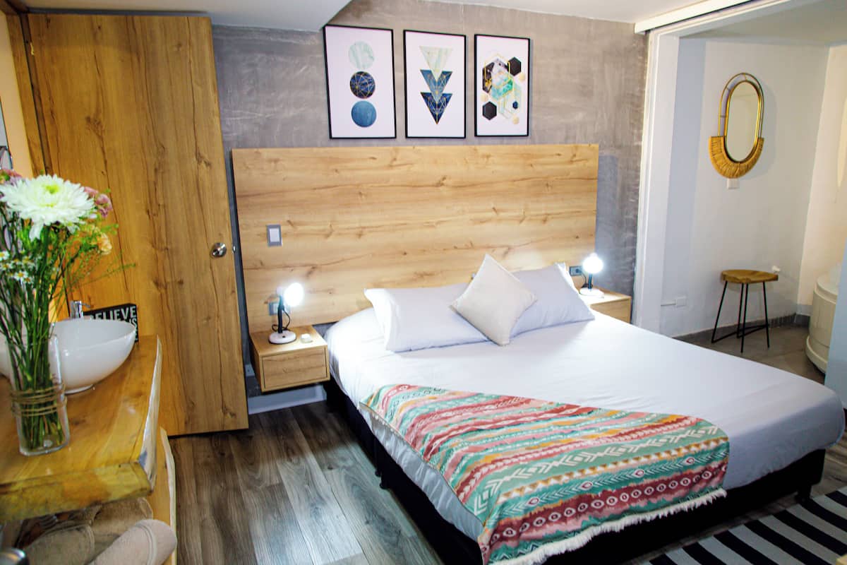 A bedroom with a wooden headboard, a bed with a colorful blanket, a bedside table with a lamp, and a wall-mounted mirror.