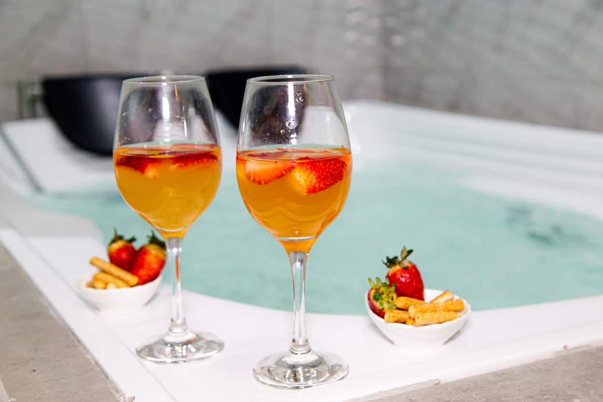 Two glasses of a yellow beverage with strawberries, a bowl of strawberries, and a bowl of fries on a white surface near a bathtub.