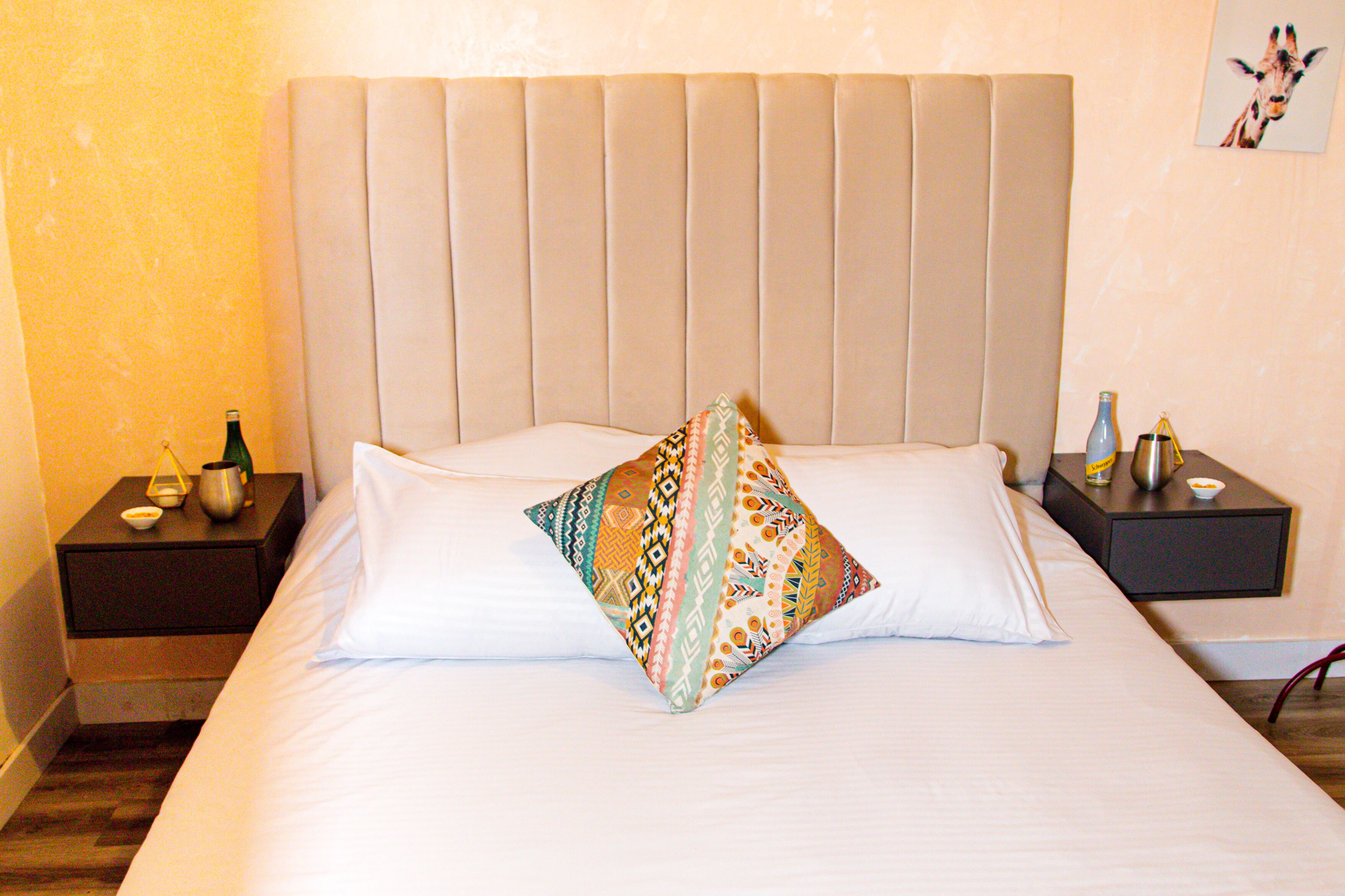 A neatly made bed with a patterned pillow, a bedside table with a bottle and a small bowl, and a nightstand with a lamp.