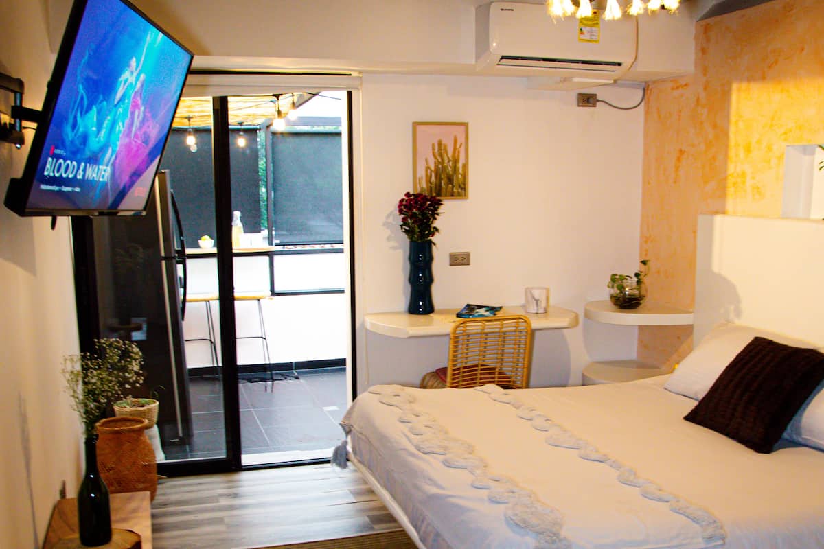 A hotel room with a bed, a TV, a desk, a chair, a small table, a vase with flowers, and a window.