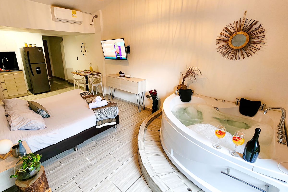 A modern hotel room with a bed, a sitting area, a TV, a bathroom with a bathtub, and a dining area.