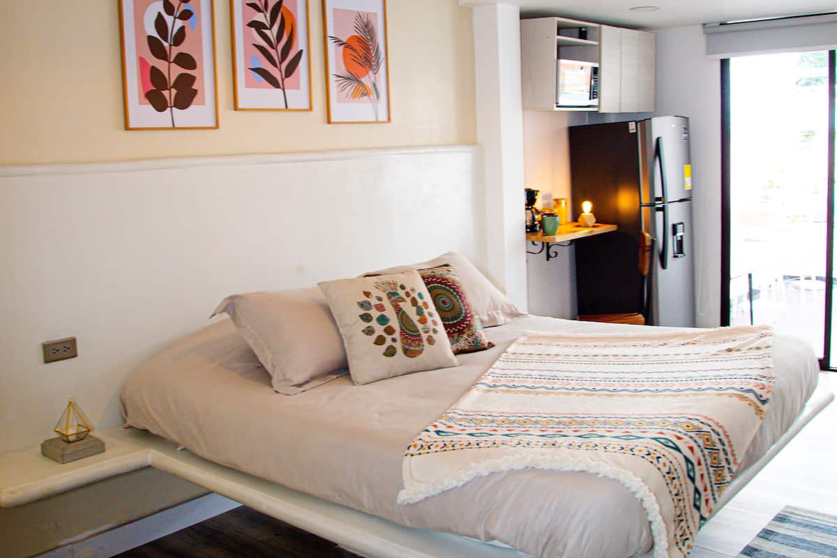 A bedroom with a bed, pillows, a bedside table, a wall-mounted shelf with a lamp, and a refrigerator.