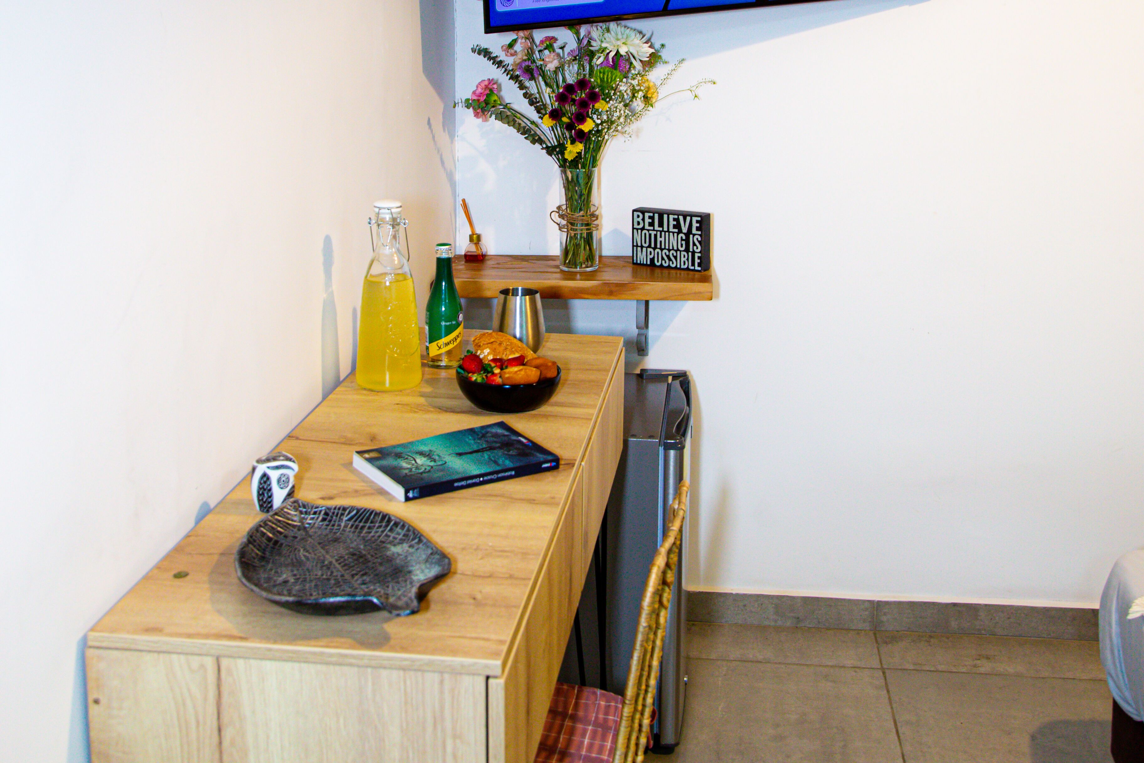 A kitchen counter with a bottle of yellow liquid, a loaf of bread, a bowl of fruit, a book, and a decorative sign.