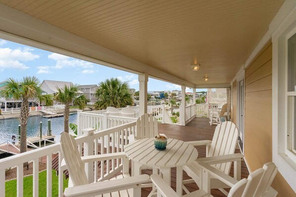 A covered outdoor deck with white chairs and a table, overlooking a body of water and residential buildings.