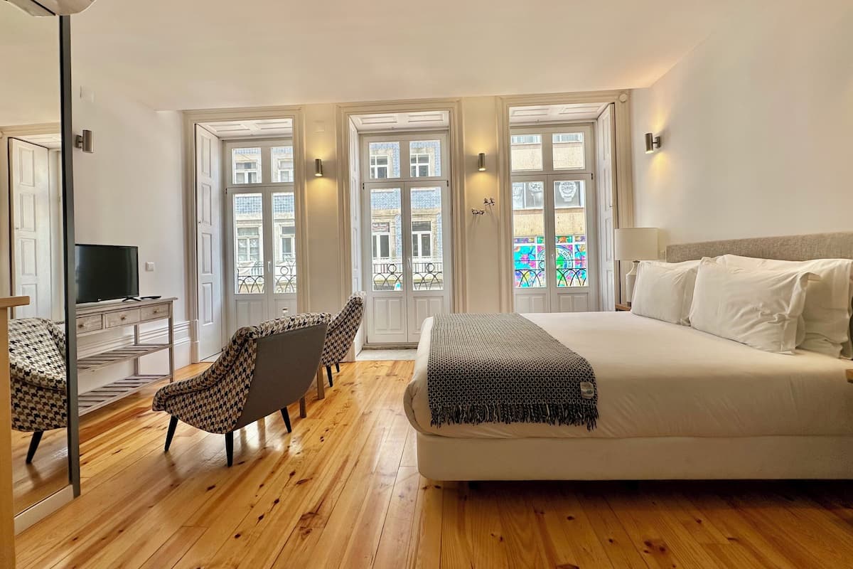 A bedroom with a bed, a desk with a TV, two armchairs, and a large window with a view of buildings.