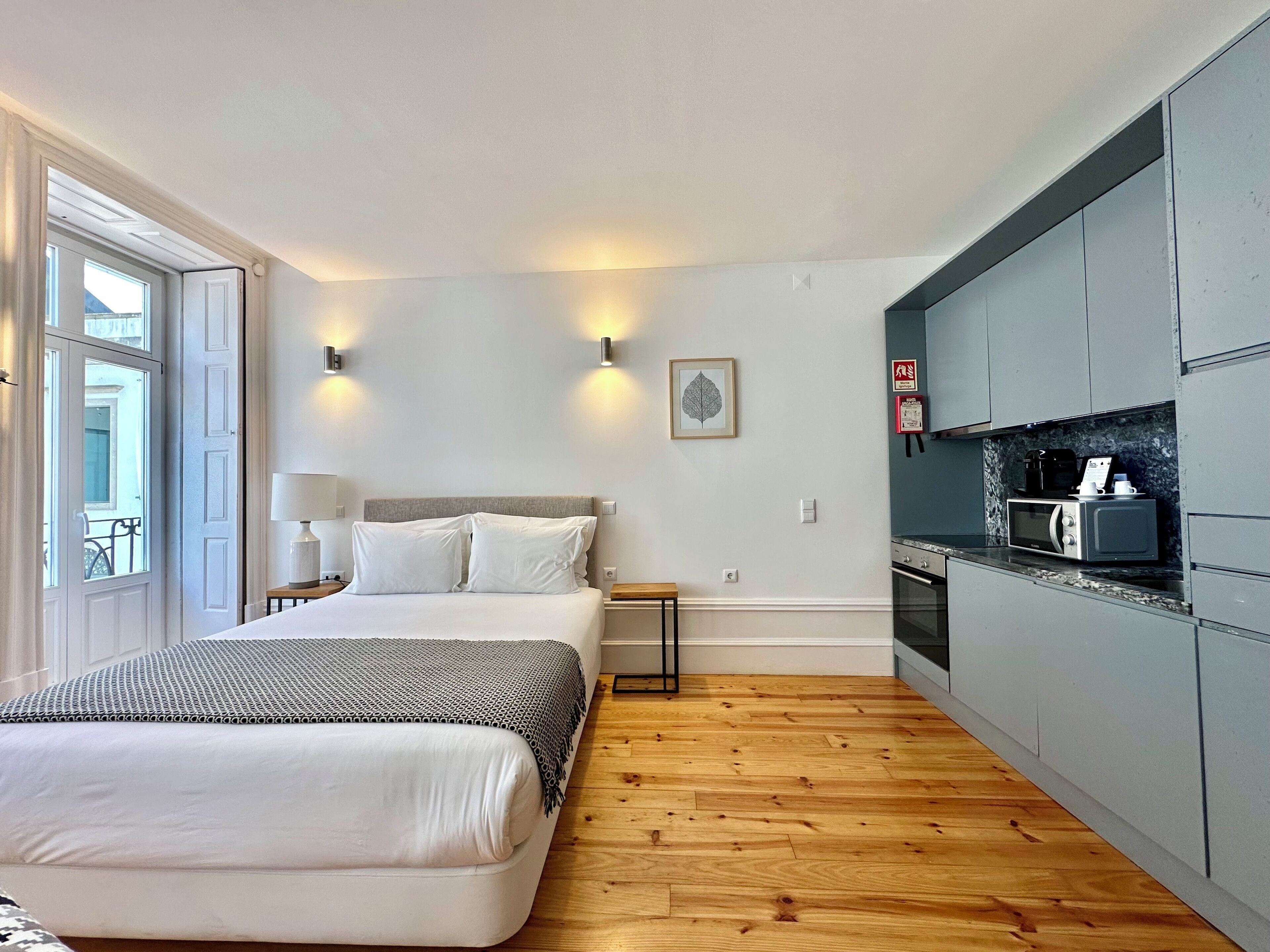 A hotel room with a bed, bedside table, lamp, wall-mounted light, framed artwork, and a kitchenette with a microwave and coffee maker.