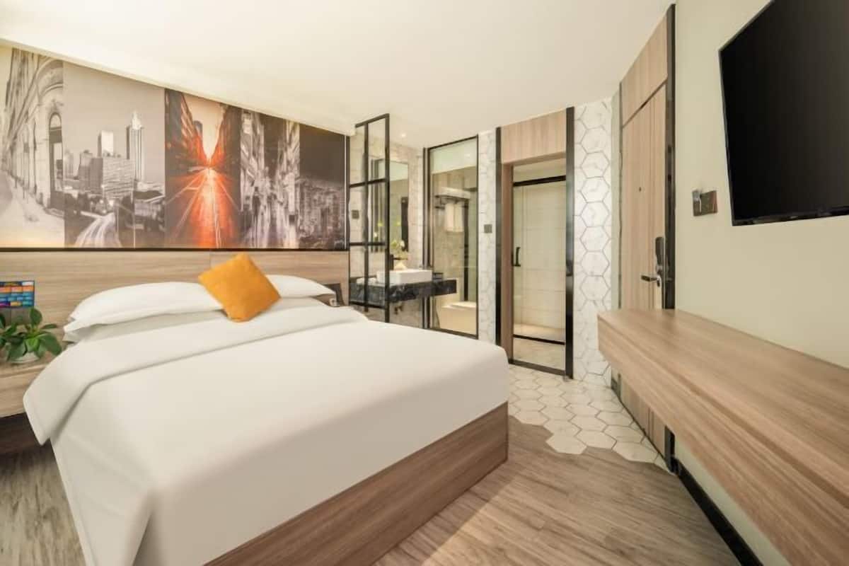 Una habitación de hotel moderna con una cama grande, televisión, baño con puerta de vidrio y un mural con vista a la ciudad.