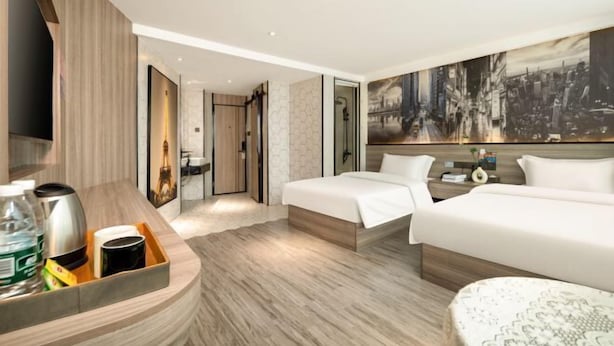 Une chambre d’hôtel avec deux lits, un grand tableau représentant un paysage urbain, un bureau en bois avec un plateau contenant divers objets et un téléviseur.