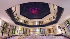L’intérieur d’un hôtel doté d’un atrium central, d’un plafond orné de motifs évoquant une galaxie et d’un espace piscine agrémenté de compositions florales.